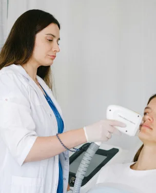 Esthetician in a white coat using a skin-analysis device on a client's face.