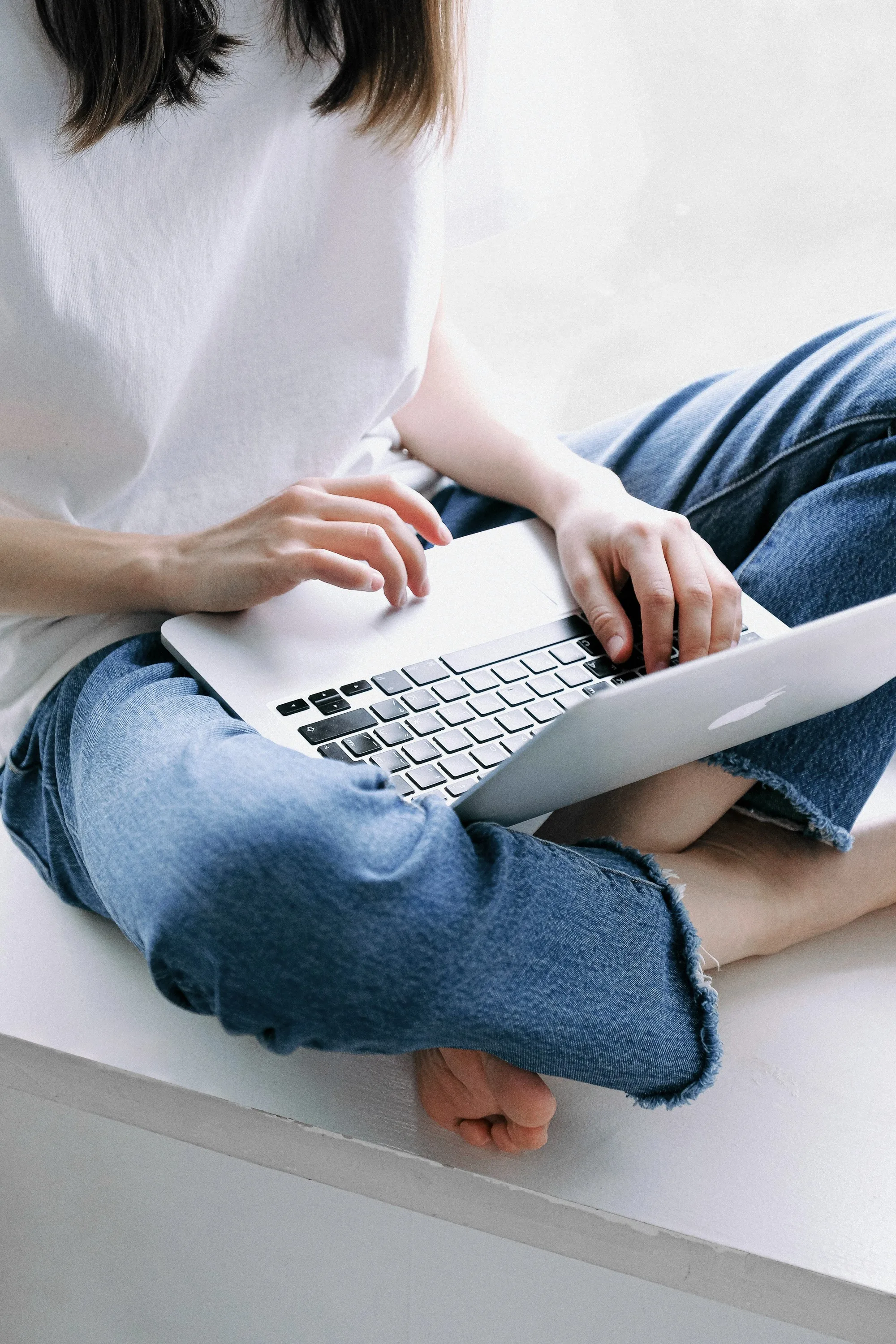 Person sitting cross-legged in jeans, working on a laptop.